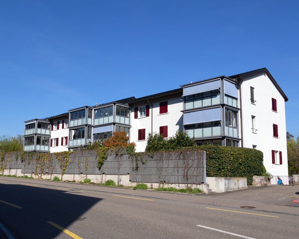 Aussenansicht der Liegenschaft von der Strasse her. Begrünte Lärmschutzmauer, helle Fassade, rote Läden, bläuliche Wintergärten.